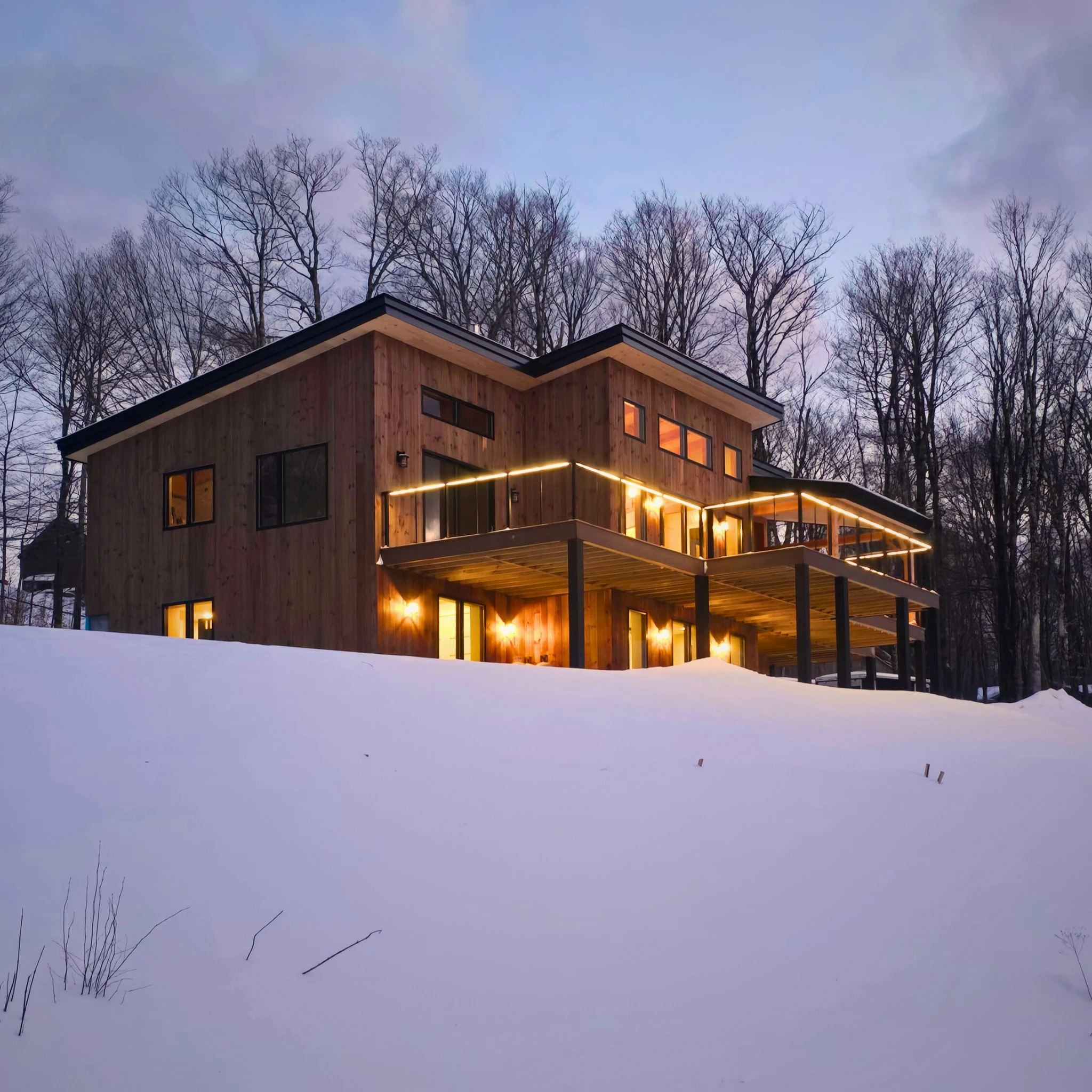 Warm interior and porch lighting on a wood clad mountain home surrounded by snow at dusk.