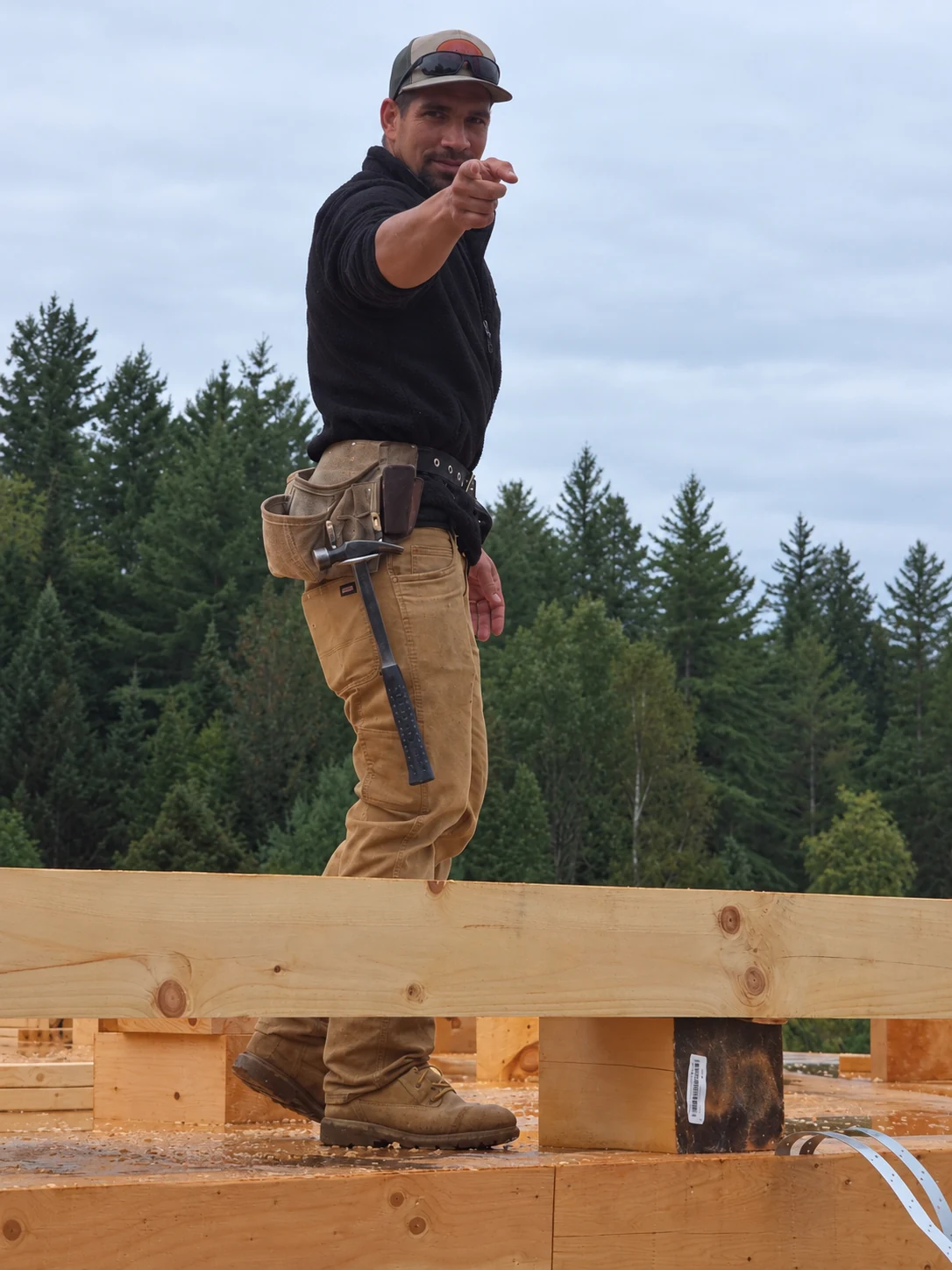 A Hudson carpenter in work boots and a toolbelt, smiling and pointing toward the camera on a timber frame jobsite.