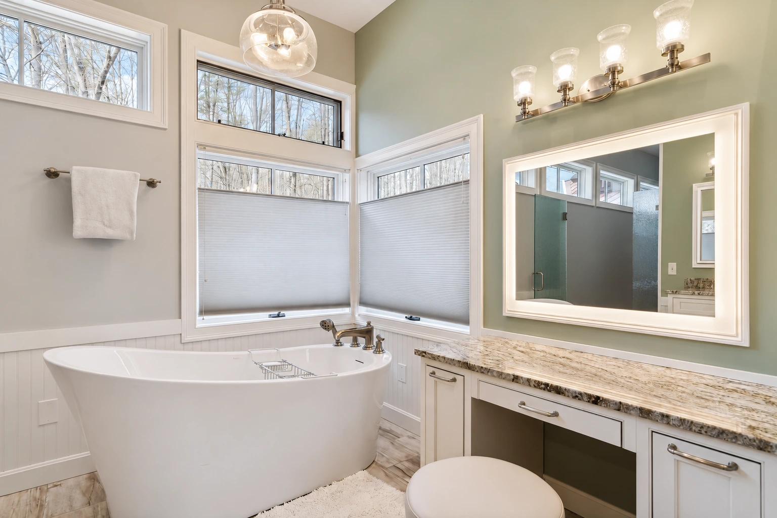 Freestanding white bathtub beneath high windows beside a pale vanity and illuminated mirror.