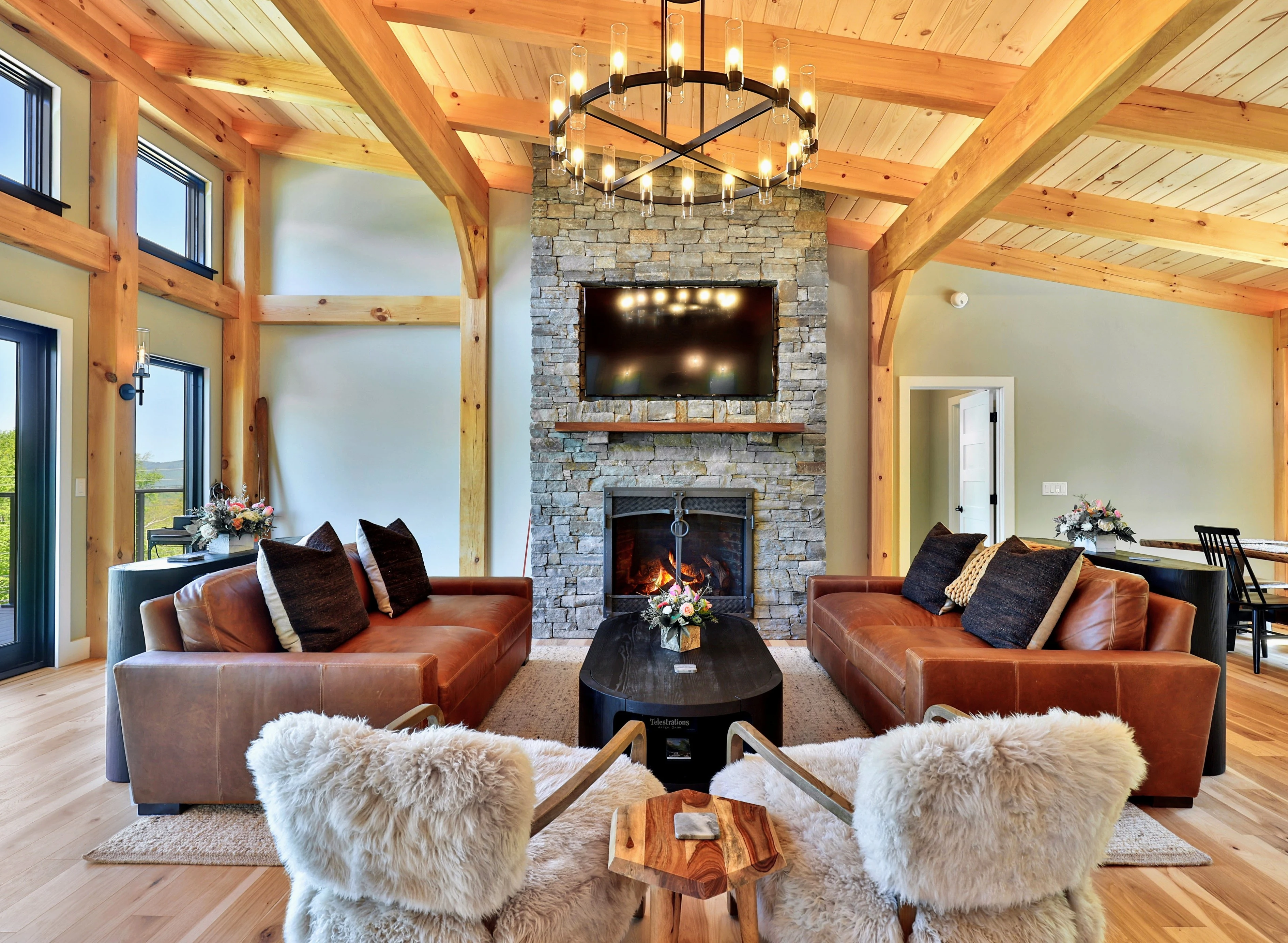 Timber frame living room with a lit stone fireplace, leather sofas and sheepskin-covered chairs in a Hudson home in Killington, Vermont.