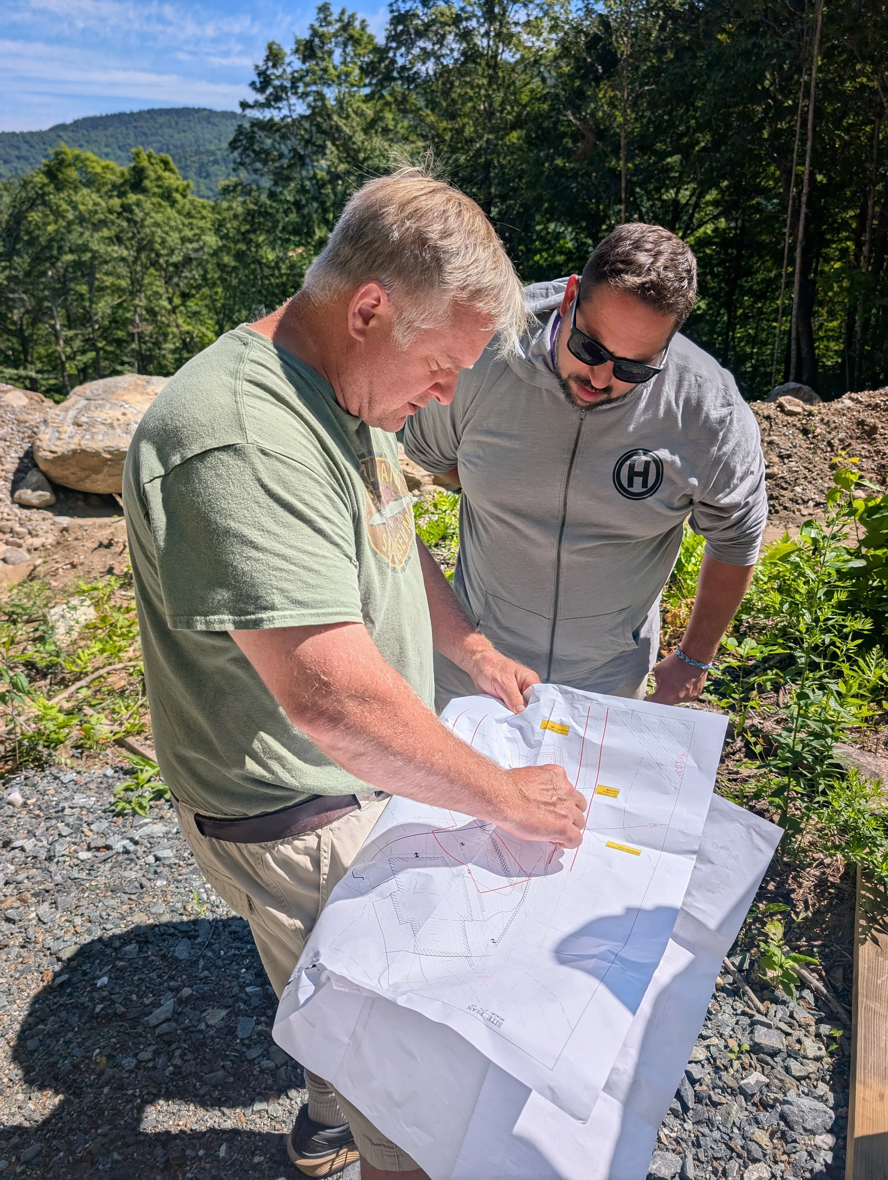 Two people reviewing a drawing together outdoors at a wooded building site.