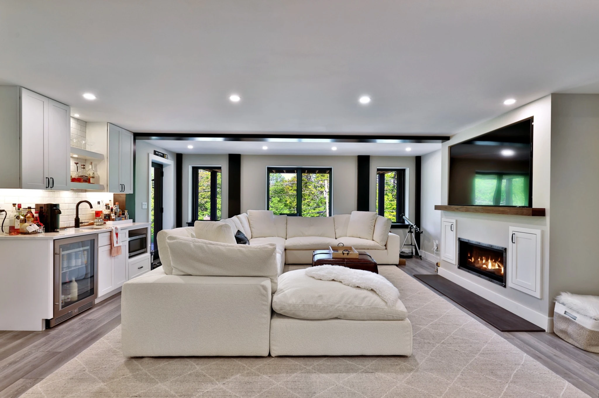 A lower-level Killington lounge with cream sectional sofas, a white wet bar, a fireplace and windows facing the trees.
