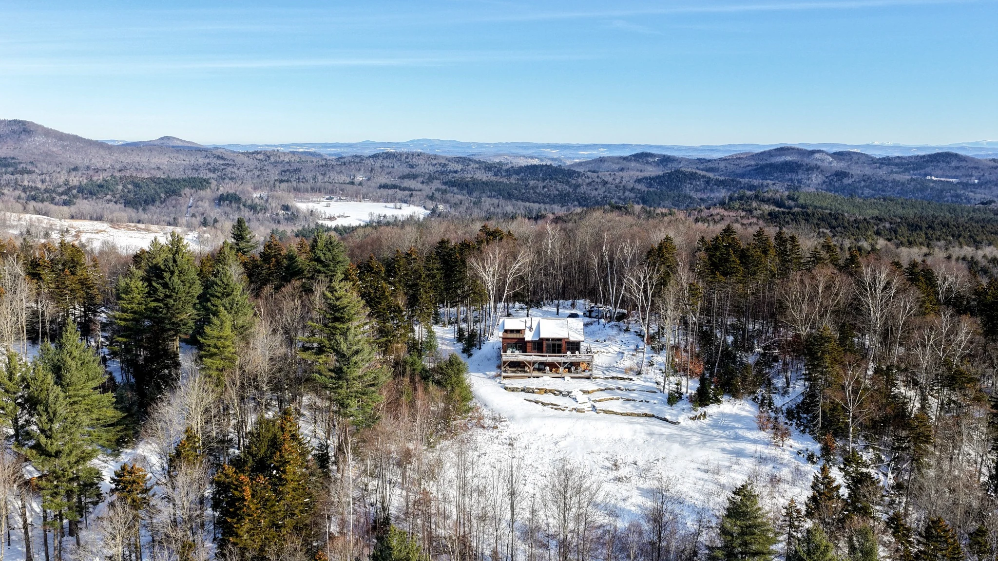 Aerial view of a snow-covered home and clearing within wooded Vermont hills, with distant mountain ridges.