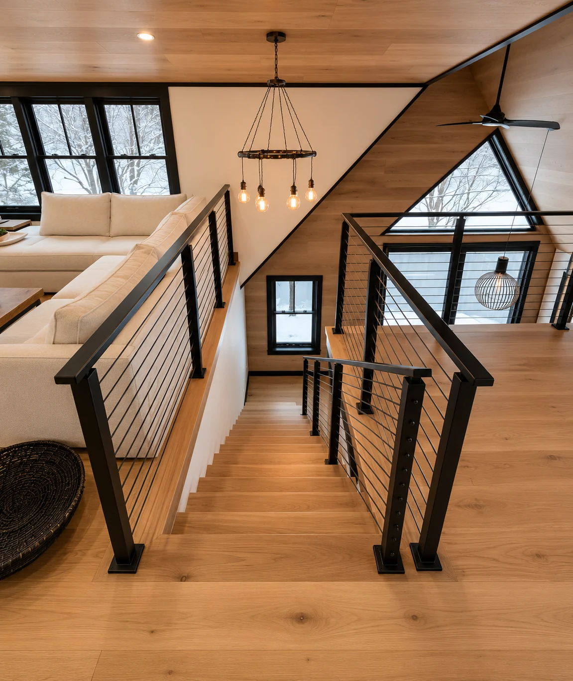 View down a wood staircase from the upper landing, with black railings and a tall triangular window beyond.