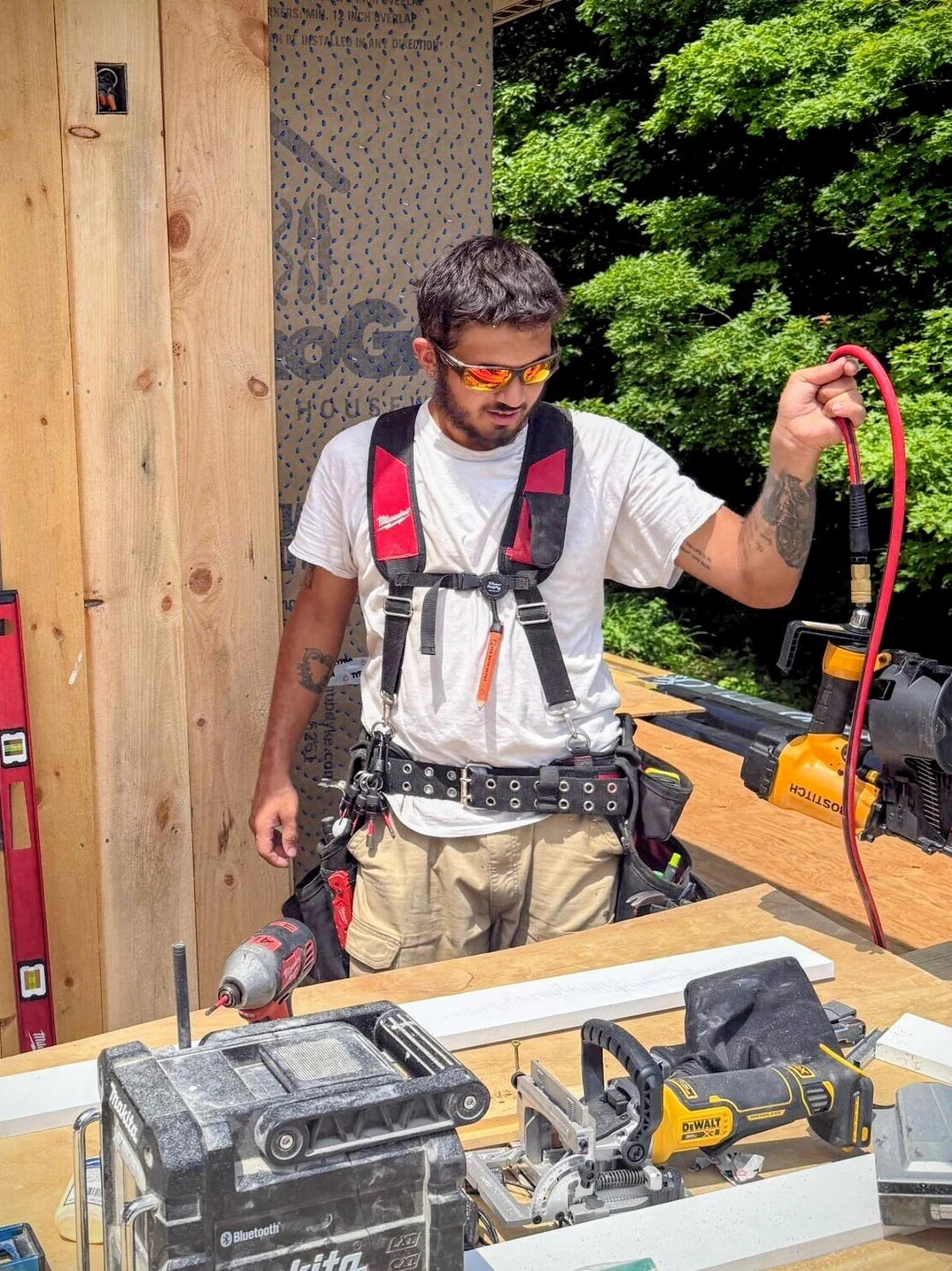 Hudson builder wearing safety glasses and a tool belt at a workbench beside timber framing.