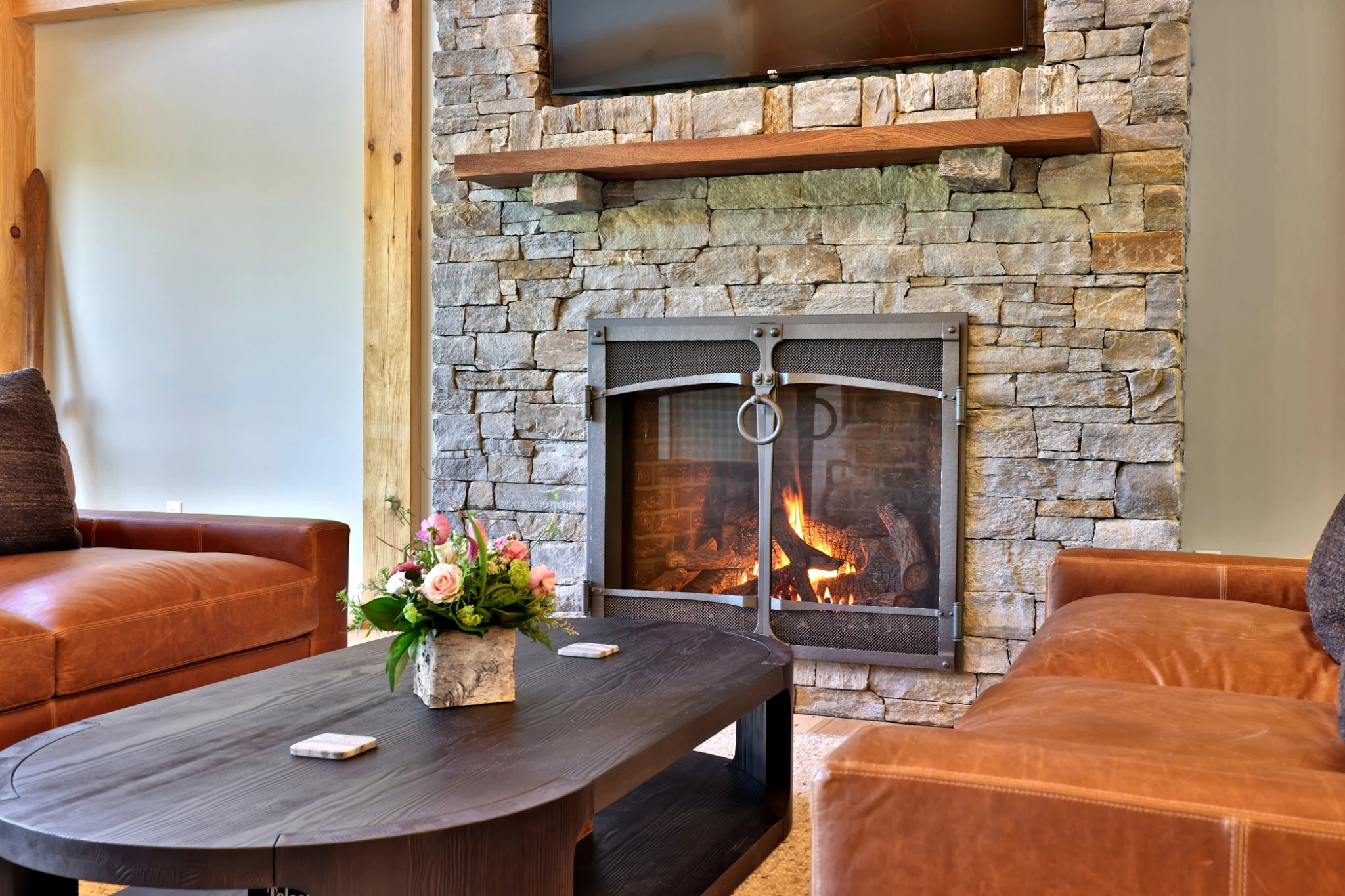 A lit stone fireplace, wood mantel and warm leather seating in the Killington timber frame home.