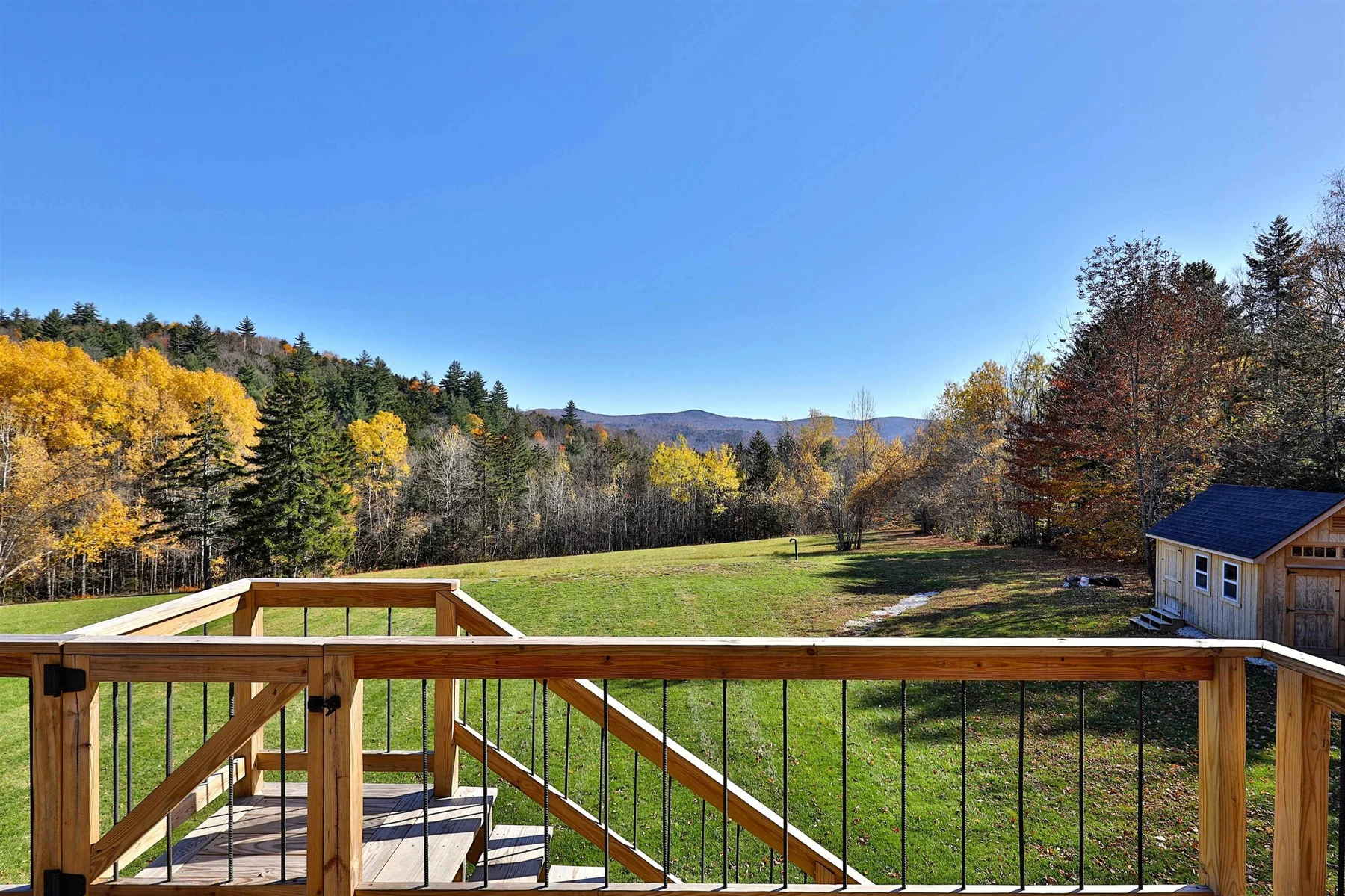 A wood deck and railing overlooking a green lawn and a wooded hillside in autumn.