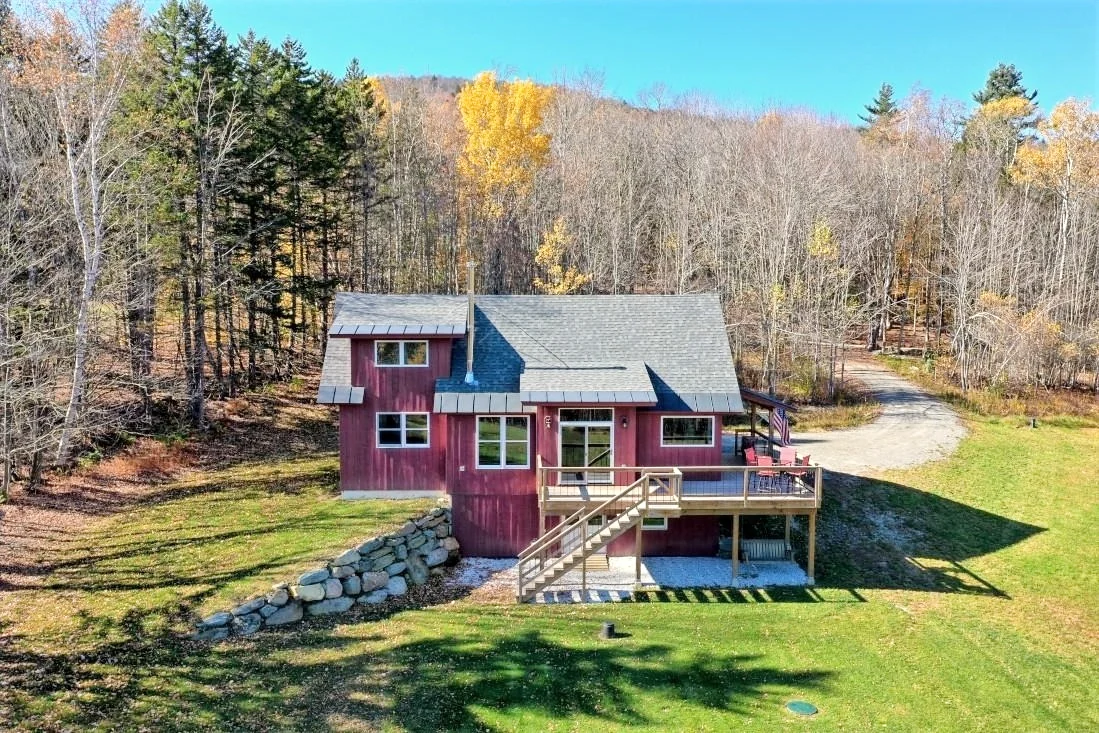 An elevated view of a red timber home, wood deck, lawn, and surrounding autumn trees.