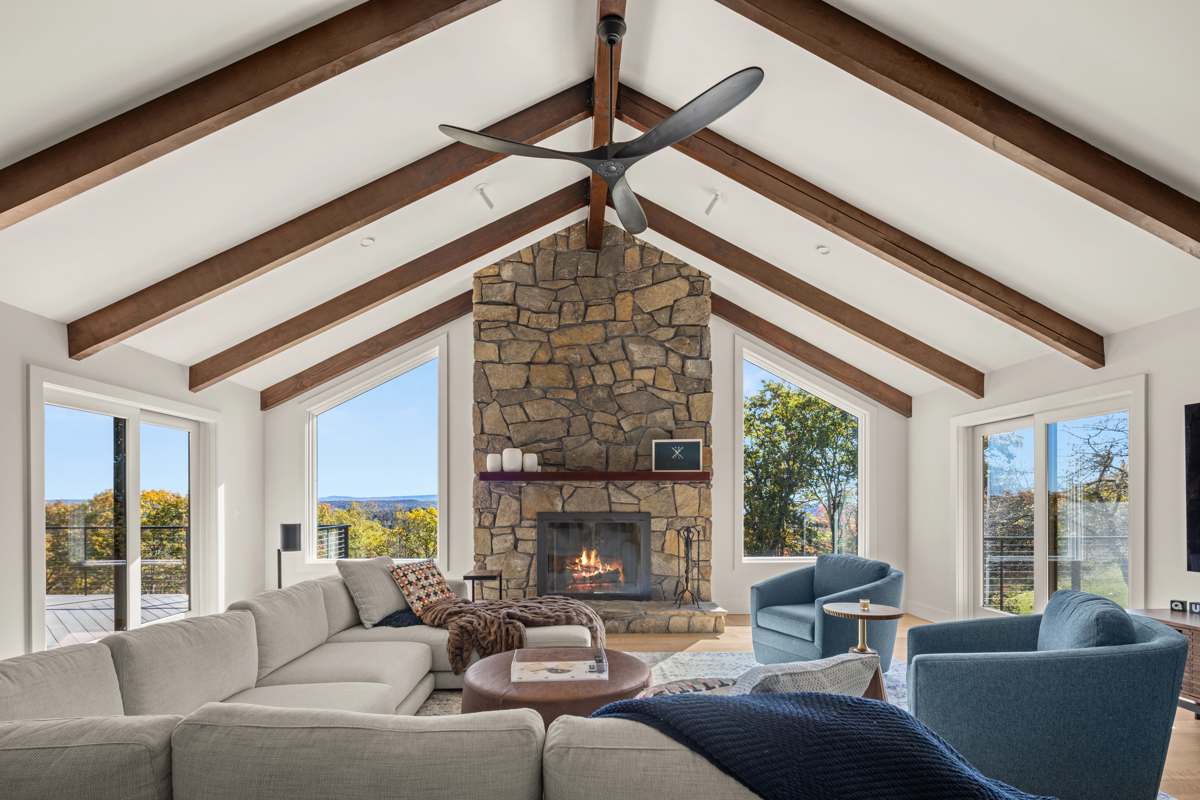 Great room with exposed beams, a stone fireplace and landscape-facing windows in Quechee.