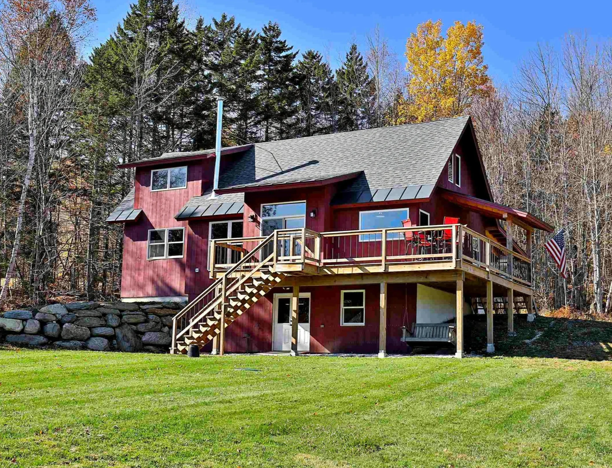 Red timber frame home with a wraparound deck and lawn beside woodland in Stockbridge, Vermont.