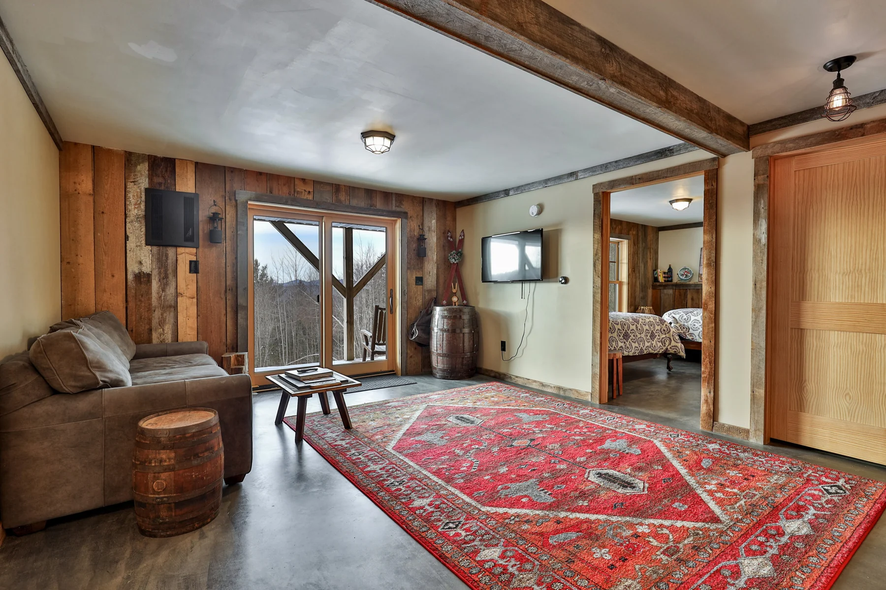 A lower-level sitting room with exposed beams, a sofa, red patterned rug, and glazed doors.