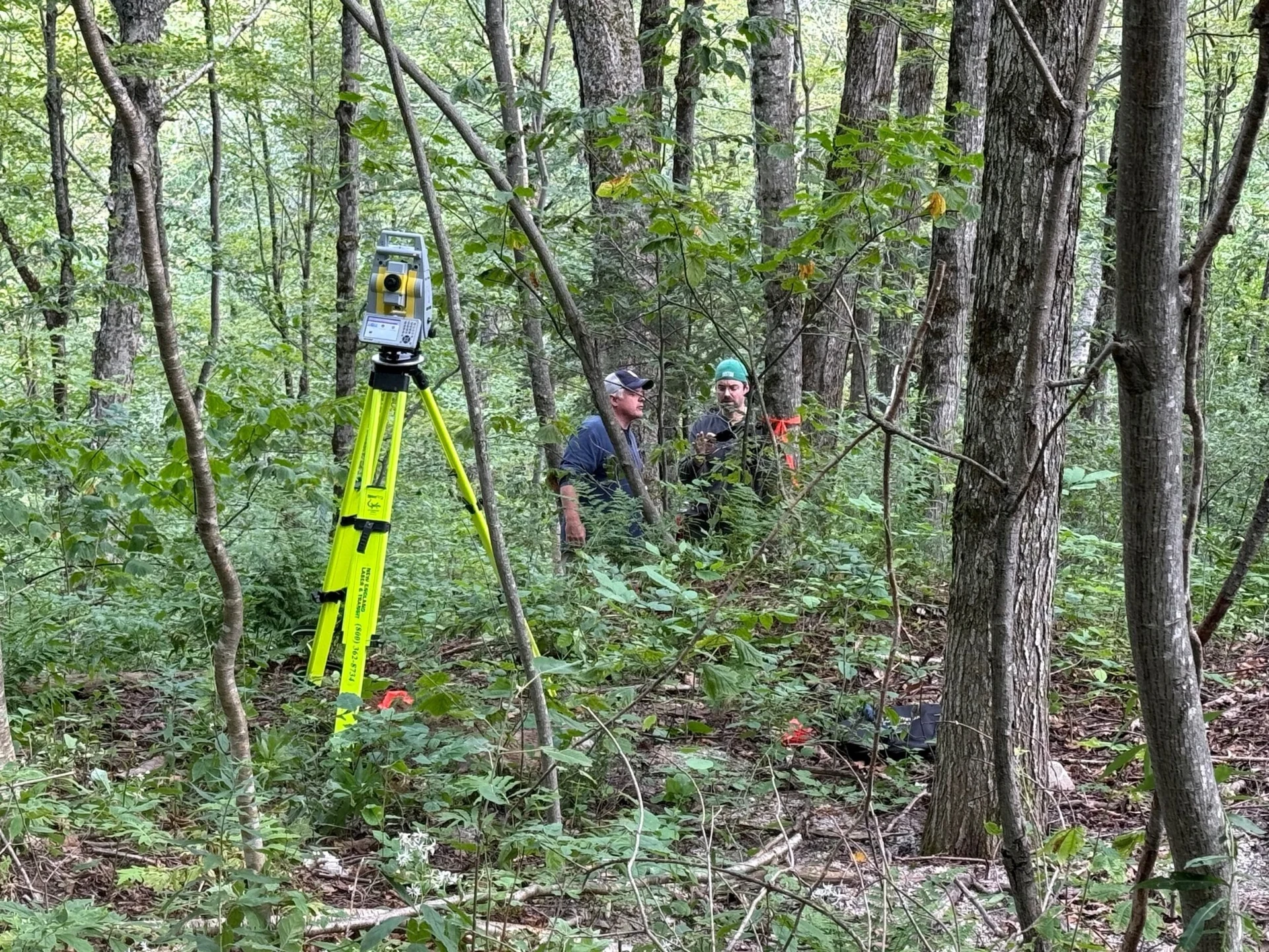 Two people working beside surveying equipment on a tripod in a wooded site.