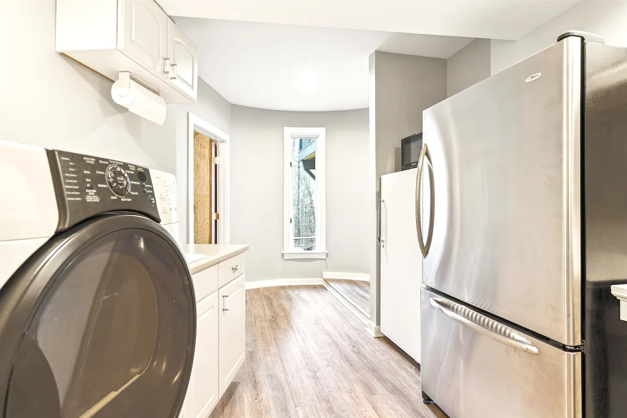 A bright laundry room with front-loading appliances, pale cabinets, and wood-look flooring.