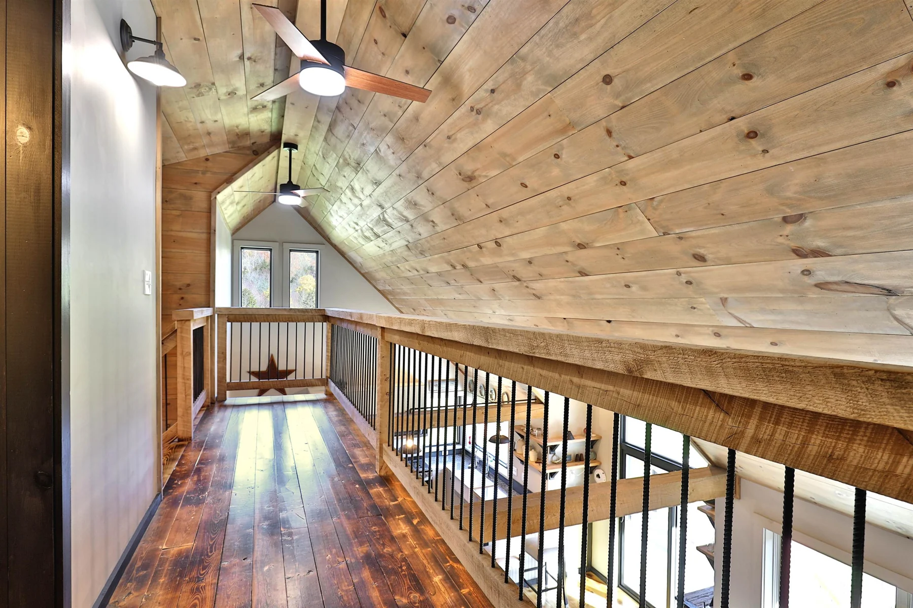 A wood-floored upper hallway with black metal railing overlooking the living space beneath a sloped wood ceiling.