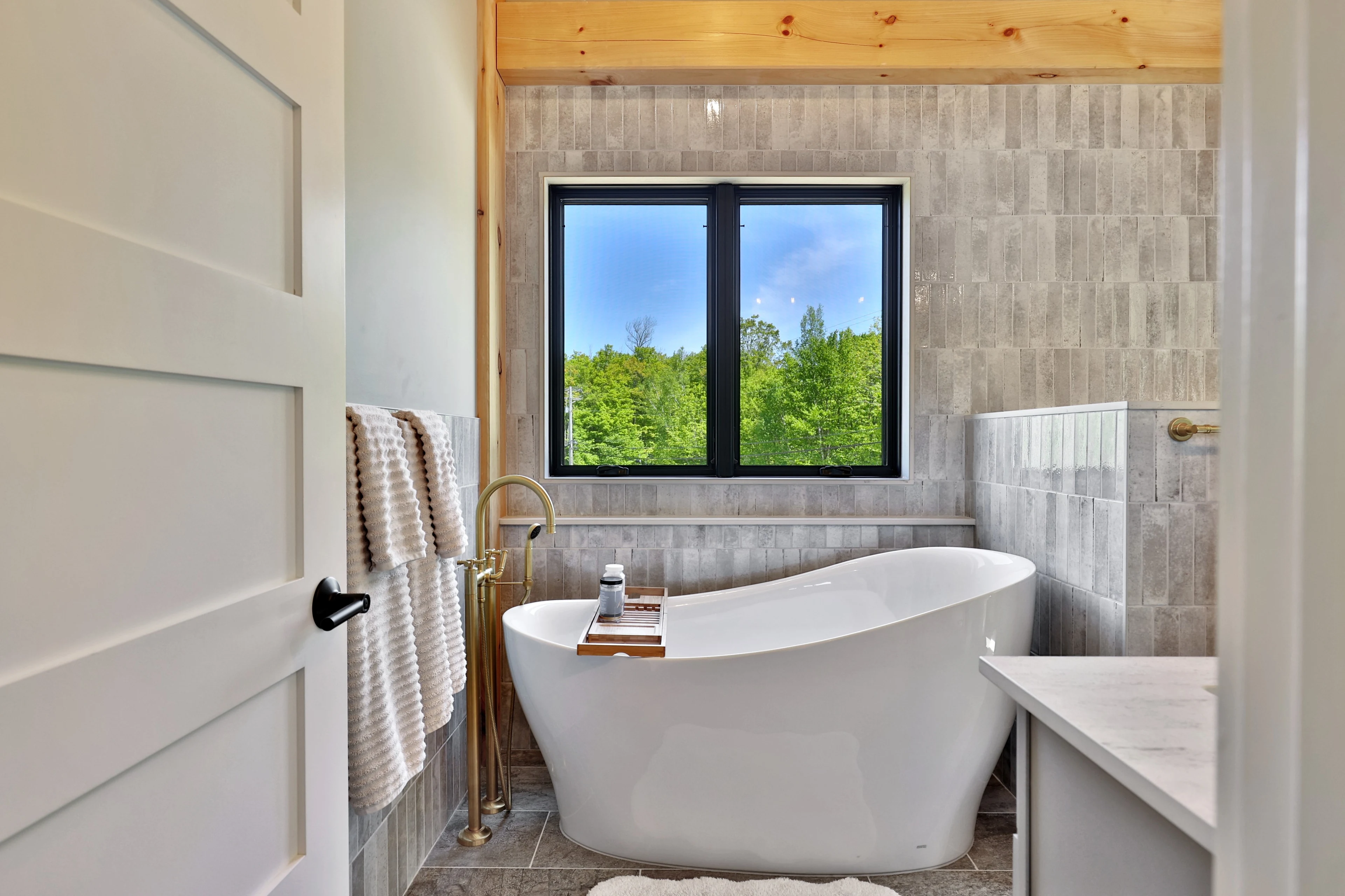 Freestanding white tub, pale tiled wall and a black-framed woodland-facing window in a Killington bathroom.