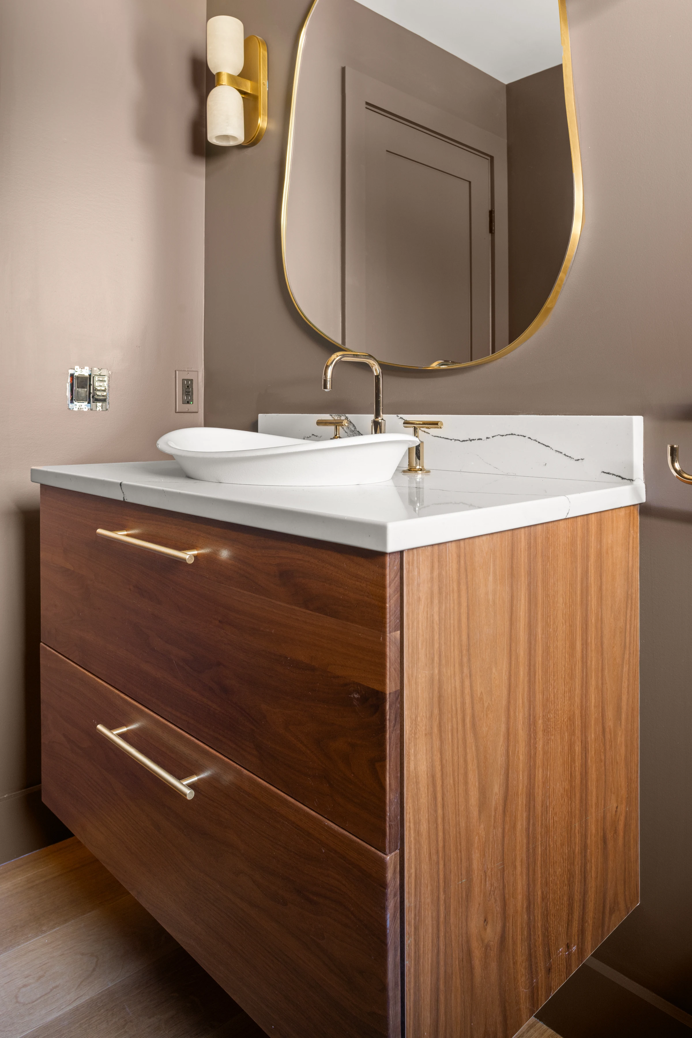 Wood vanity with a white vessel sink, veined countertop and brass-framed oval mirror.