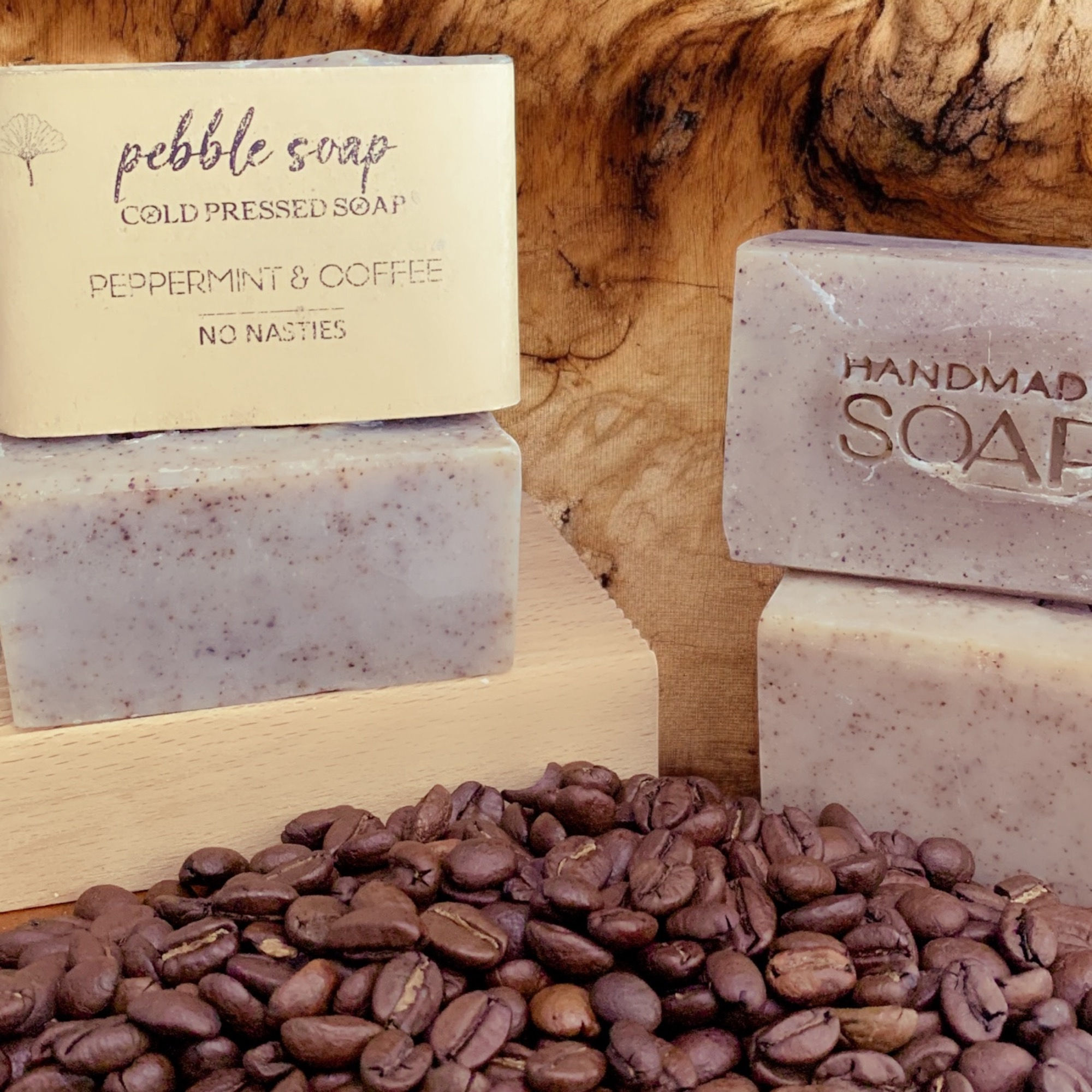 Coffee beans in the foreground 4 rectangular soaps stacked in 2 piles, one soap has a label soaps have brown flecks
