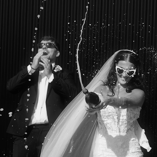 black & white photo of bride & groom laughing while shaking a champagne bottle, spraying the drink everywhere