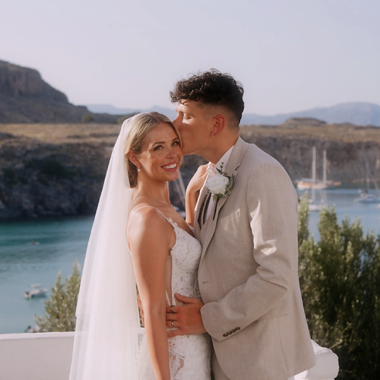 bride looking towards camera, groom kissing forehead, Greek water cove, sun