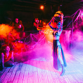 Lady Corbeau performing in a strappy black outfit on a brightly illuminated stage with smoke.