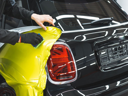 Someone places a bright yellow vinyl on the side of a black car. They are using a tool to smooth the wrap over the vehicle.