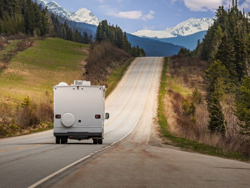 A white RV driving along a two-lane road between evergreen forests and grassy hills toward snowy mountain peaks.