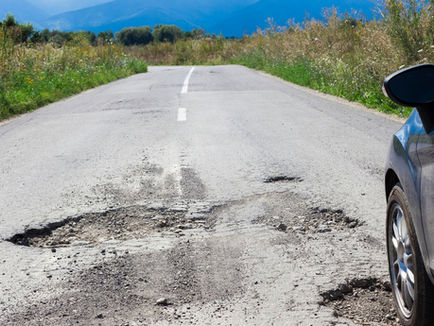 A back road with a car driving over potholes in the asphalt. A field of wild vegetation is on either side of the road.