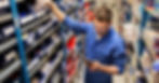 An employee taking a bag of car parts down from a shelf in an auto repair shop. The employee holds a tablet.