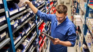 An employee taking a bag of car parts down from a shelf in an auto repair shop. The employee holds a tablet.