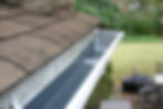 A close-up of a clean, freshly installed house gutter along a roof with brown shingles. Th