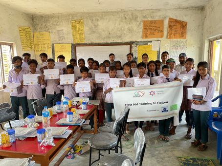 教室で制服の生徒たちが修了証を掲げ、ASHA/KARMA HEALTHの「First Aid Training in Rajpur」横断幕を持つ集合写真。
