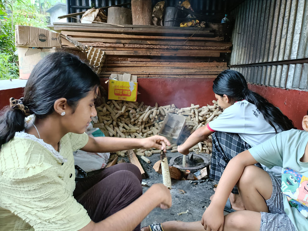 小屋の中で、3人の少女が火鉢の上でトウモロコシを焼いている。薪と段ボールが積まれ、素朴で和やかな雰囲気。