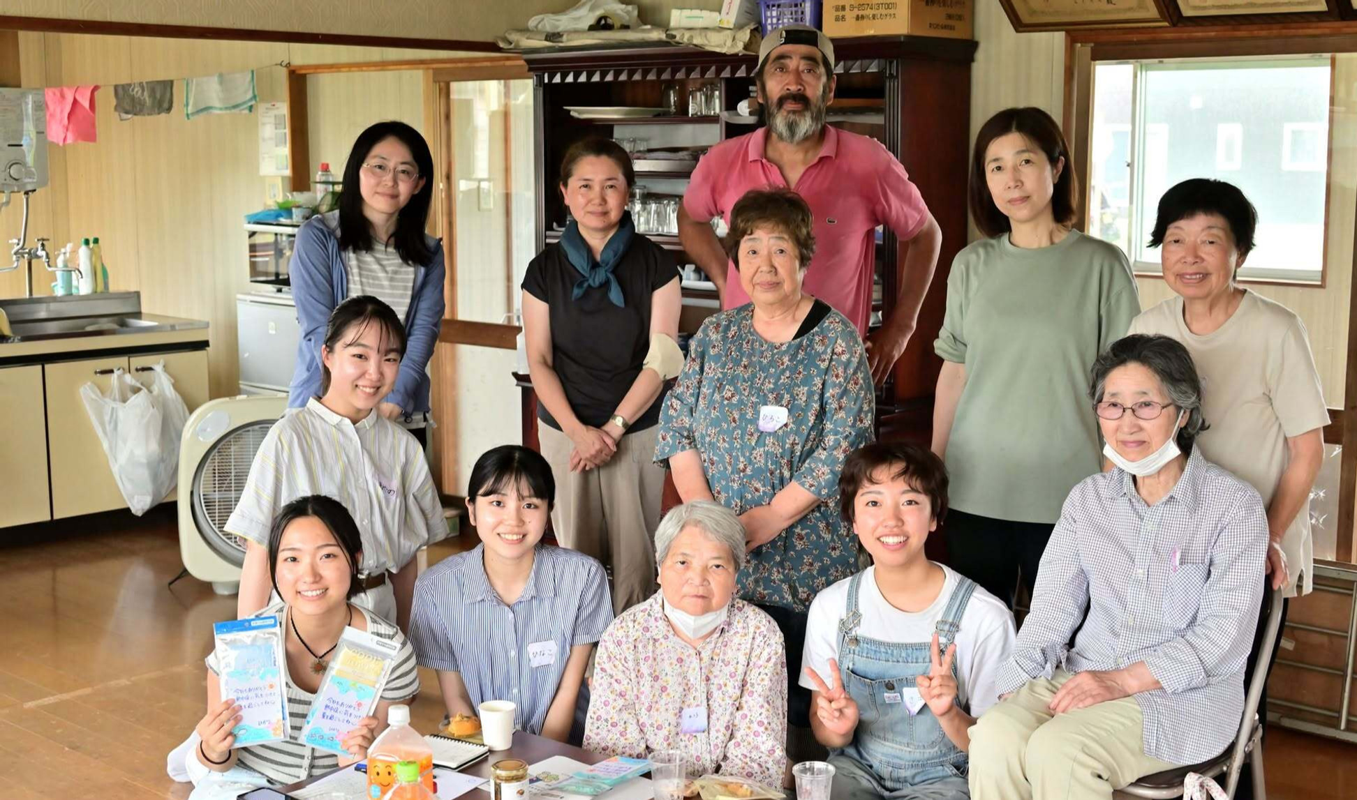 室内で年齢の異なる男女12人ほどが集合写真を撮っている。笑顔の人が多く、食卓と棚、窓が背景に見える。
