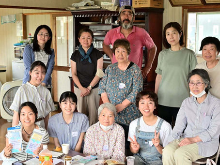 室内で年齢の異なる男女12人ほどが集合写真を撮っている。笑顔の人が多く、食卓と棚、窓が背景に見える。