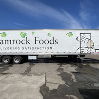 Shamrock Foods trailer with logo and text Delivering Satisfaction against blue sky.