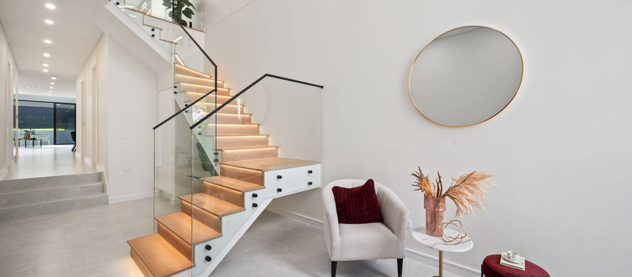 Illuminated timber staircase with glass balustrades within a contemporary double-height residential interior.