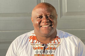 Clenny Thebe standing infront of a grey wall smiling. He is wearing an African inspired shirt that has orange elephants and grey diamonds on the middle.