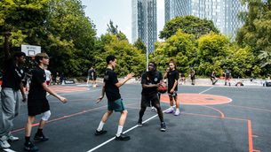 A group of young people wearing black t-shirts playing basketball. the court is black with red lines, and is surrounded by high rise buildings and trees