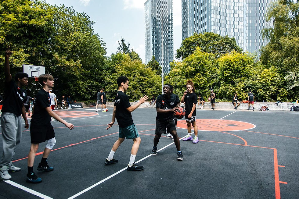 A group of young people wearing black t-shirts playing basketball. the court is black with red lines, and is surrounded by high rise buildings and trees
