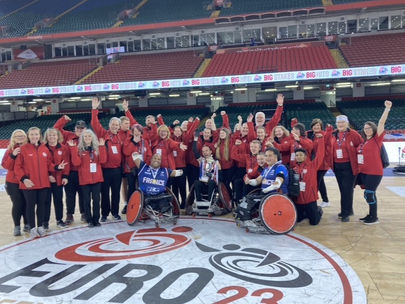 20 volunteers wearing red jackets and black trousers celebrating with Wheelchair Rugby players
