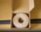Open cardboard box with a coiled rope inside, placed on a woven mat. The setting appears simple and tidy. No text visible.