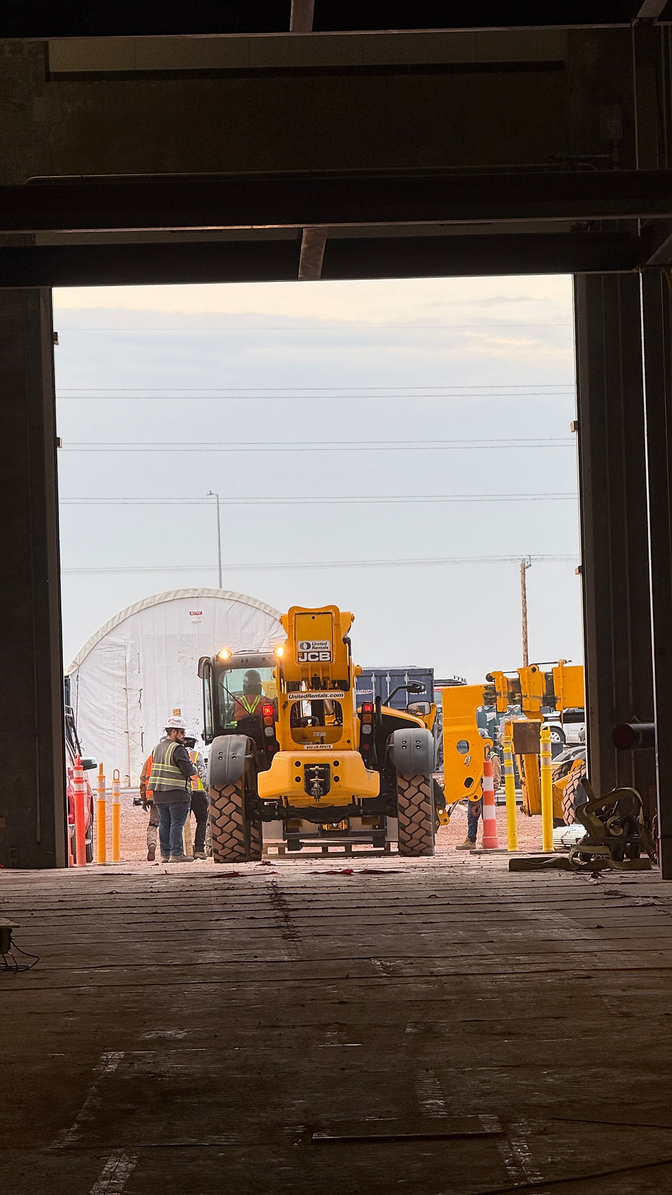 Construction site view from inside a building. A yellow JCB vehicle is centered with workers nearby. Overcast sky and industrial equipment visible.