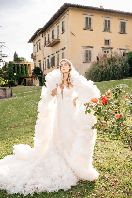 A bride in a luxurious Pronovias Privée gown with dramatic tulle layers and a jeweled headpiece poses confidently in front of a historic Tuscan villa, with vibrant red roses in the foreground. A perfect Tuscany wedding moment.