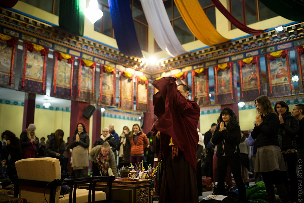 A maroon-robed teacher adjusts his robe as retreat participants stand in the temple hall — Karmapa International Buddhist Institute, New Delhi, India — December 2014 — KIBI Winter Retreat 2014 - Part 2 - The Many Smiles of Dupsing — photograph by Tokpa Korlo