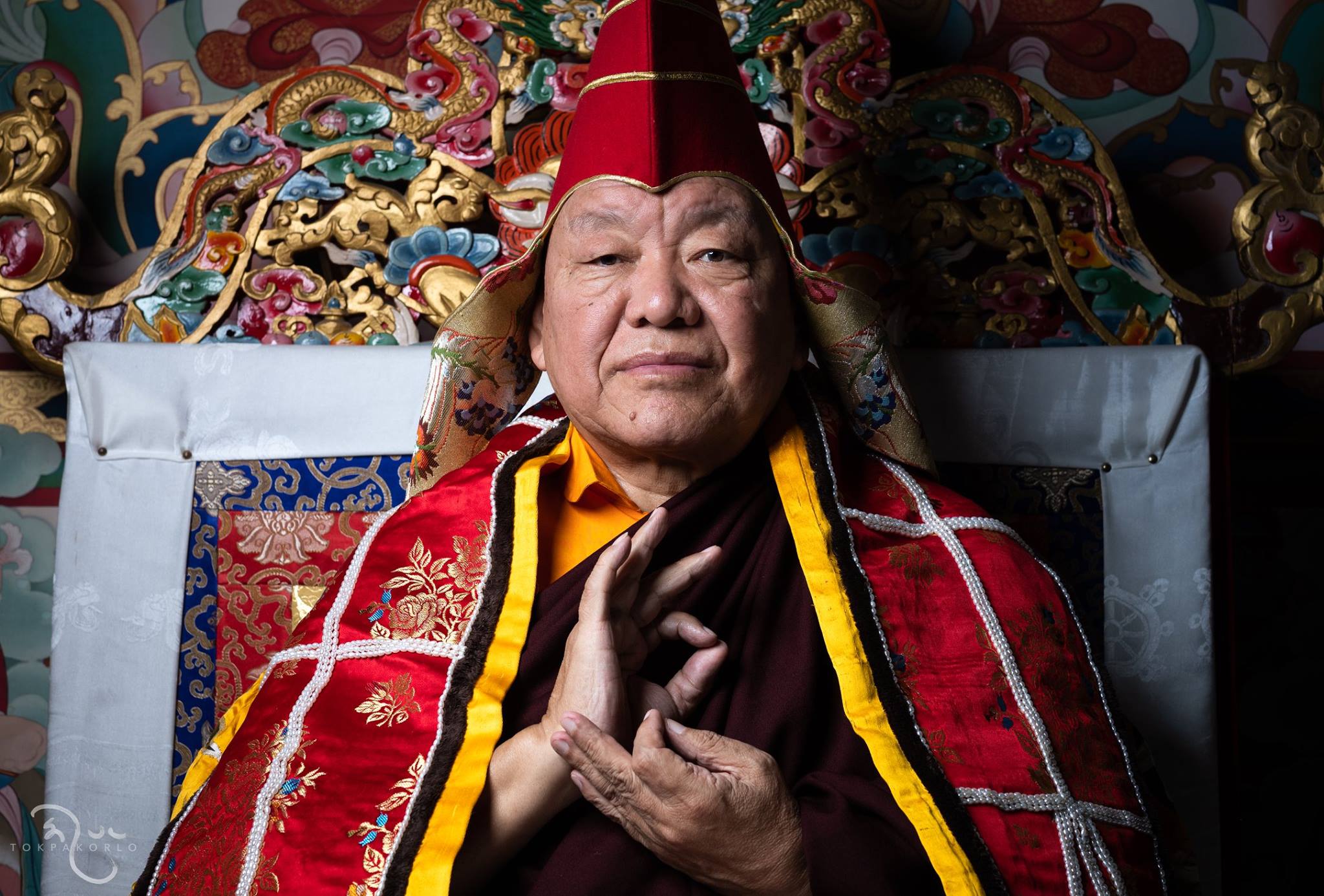 A seated figure in ornate red and gold robes gestures before a richly detailed ceremonial tapestry — The Dharma Studio — Nov 7, 2018 — The Dharma Studio - Beru Kyentse Rinpoche — photograph by Tokpa Korlo