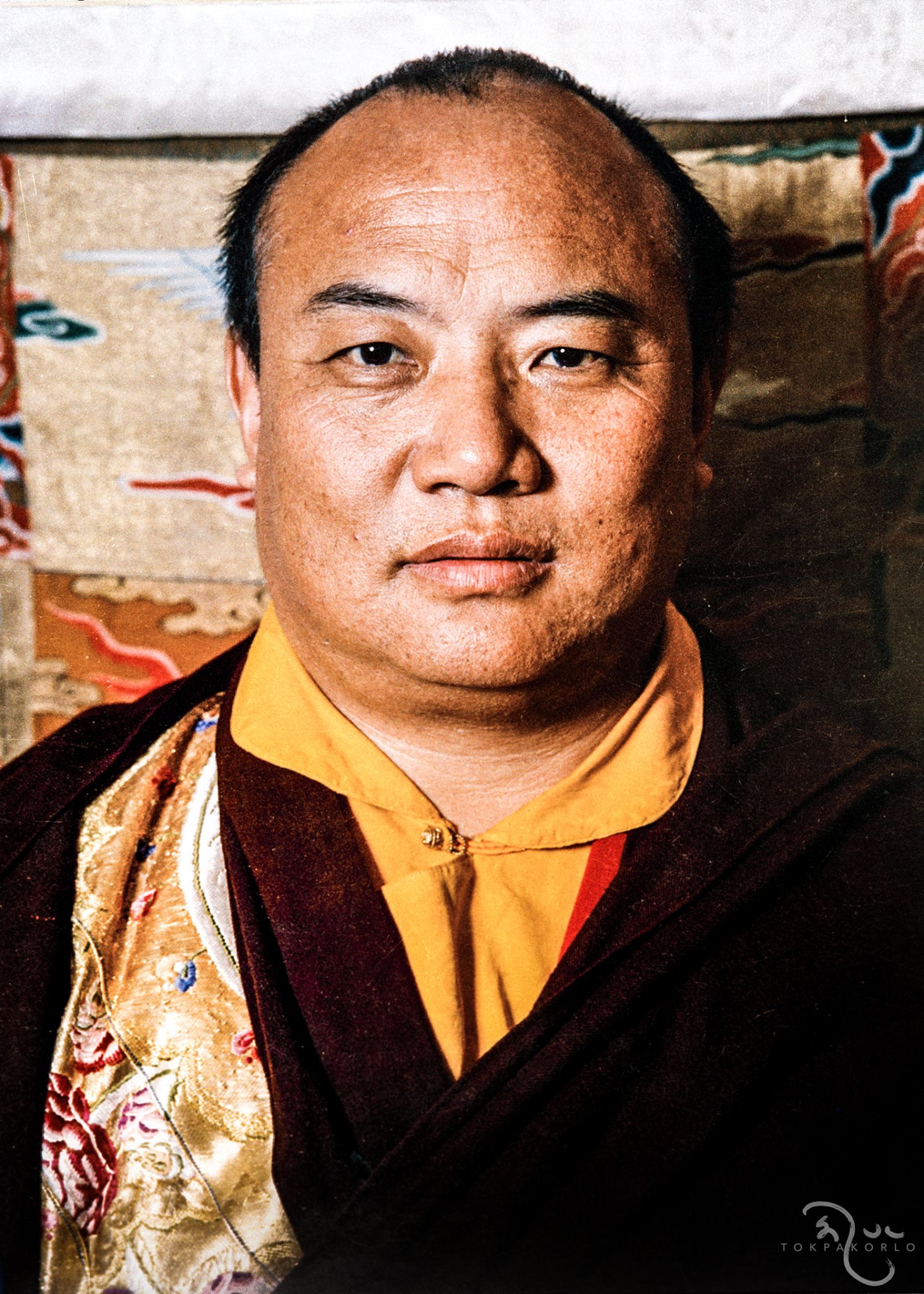 An archival color portrait of a seated figure in gold and maroon robes, facing the camera against a patterned textile — original photograph by an uncredited photographer; remastered by Tokpa Korlo — Remembering the 16th Karmapa