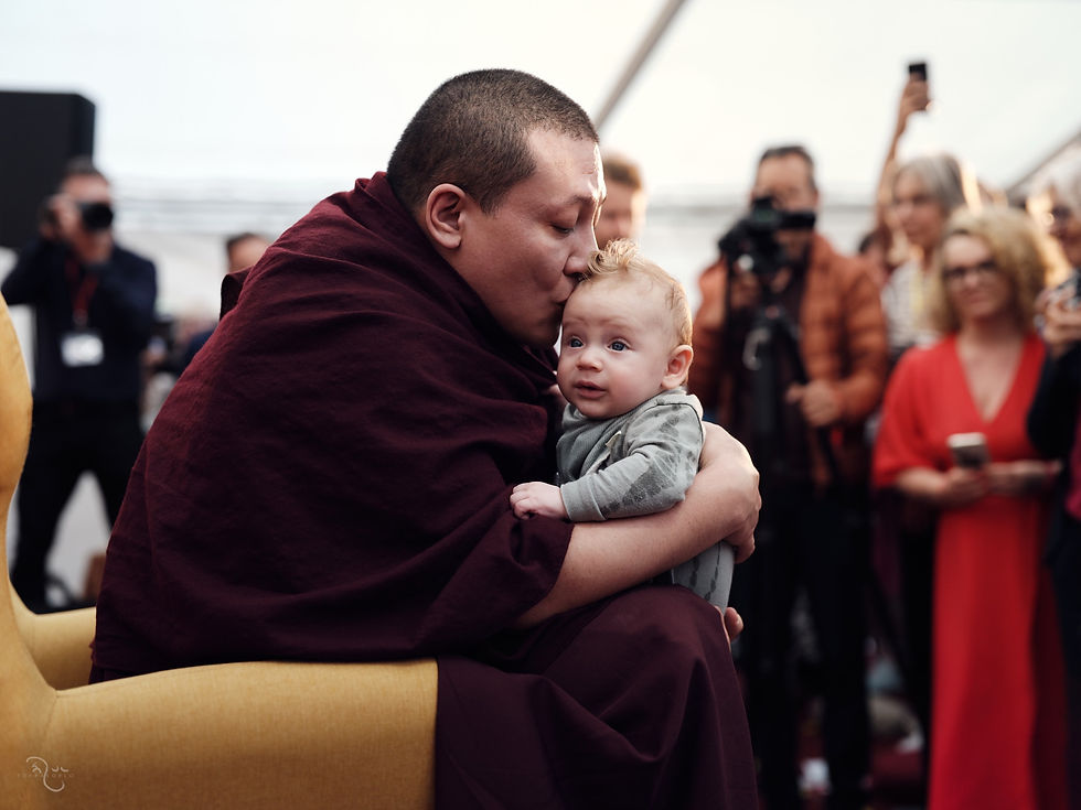 An adult in maroon robes gently cradles an infant while a small crowd watches in the background — London, United Kingdom — Jul 28, 2024 — London - Babies, Babies, Babies — photograph by Tokpa Korlo