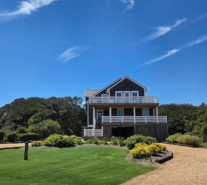Duck ocean side house with landscaped lawn including multiple flower beds, mulch, salt-tolerant plants, trees, and rock.