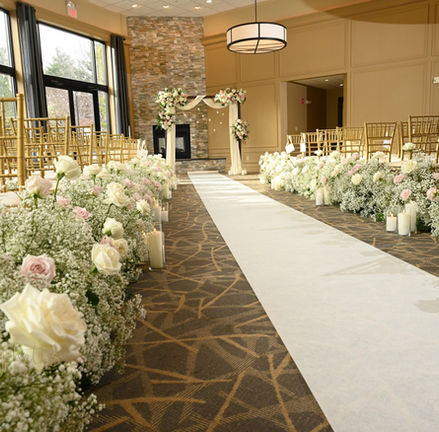 We see a think hedge of white roses and babies breath floral arrangements on either side of a white aisle runner extending from bottom right towards a large flower covered arch way at the end of the aisle at Miraval Gardens in East Bridgewater MA