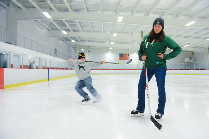 The groom is spraying the bride with ice shards from racing up behind her on the ice rink as part of this special engagement session at an ice arena on Cape Cod